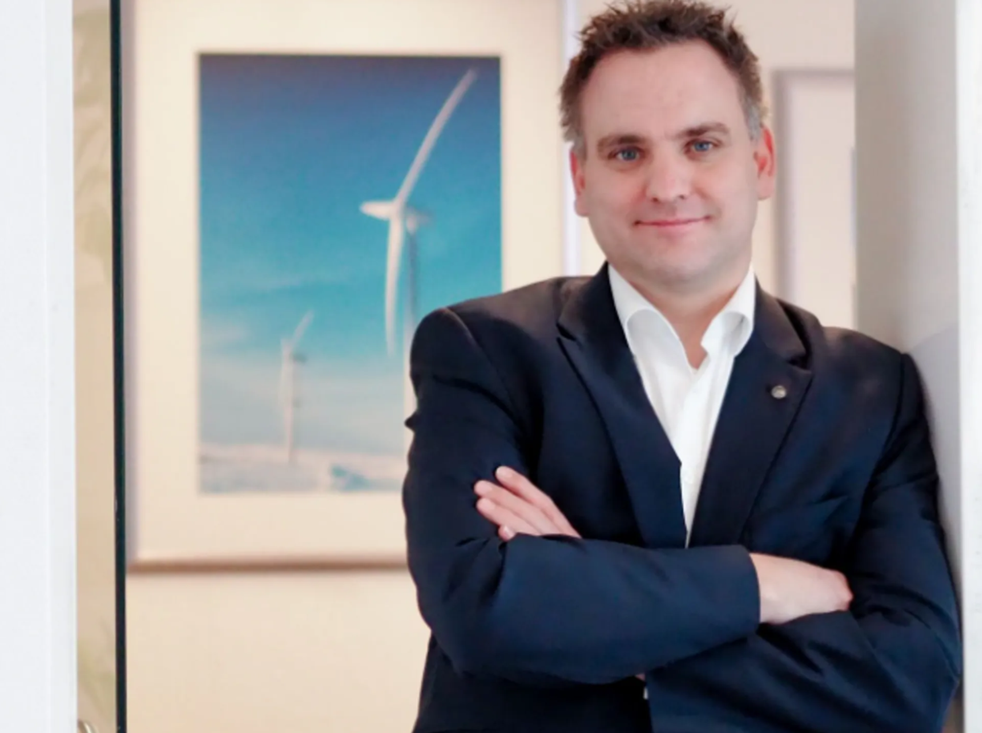 Dr.-Ing. Daniel Becker, thyssenkrupp rothe erde, wearing a dark suit with arms crossed, with a photograph of a wind turbine in the background.