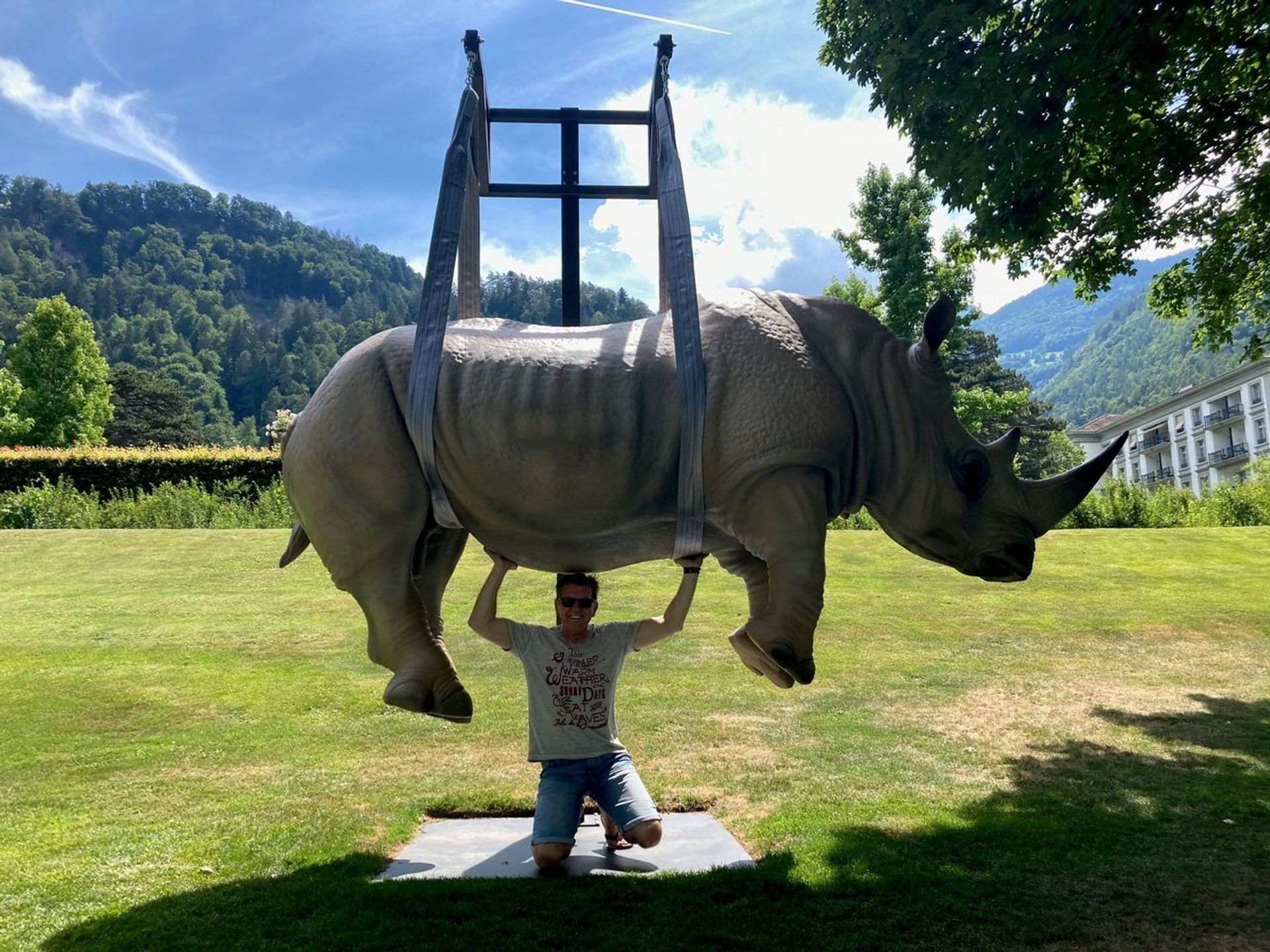 männlich gelesene Person hockt unter Statue eines Nashornes, welche er augenscheinlich stemmt
