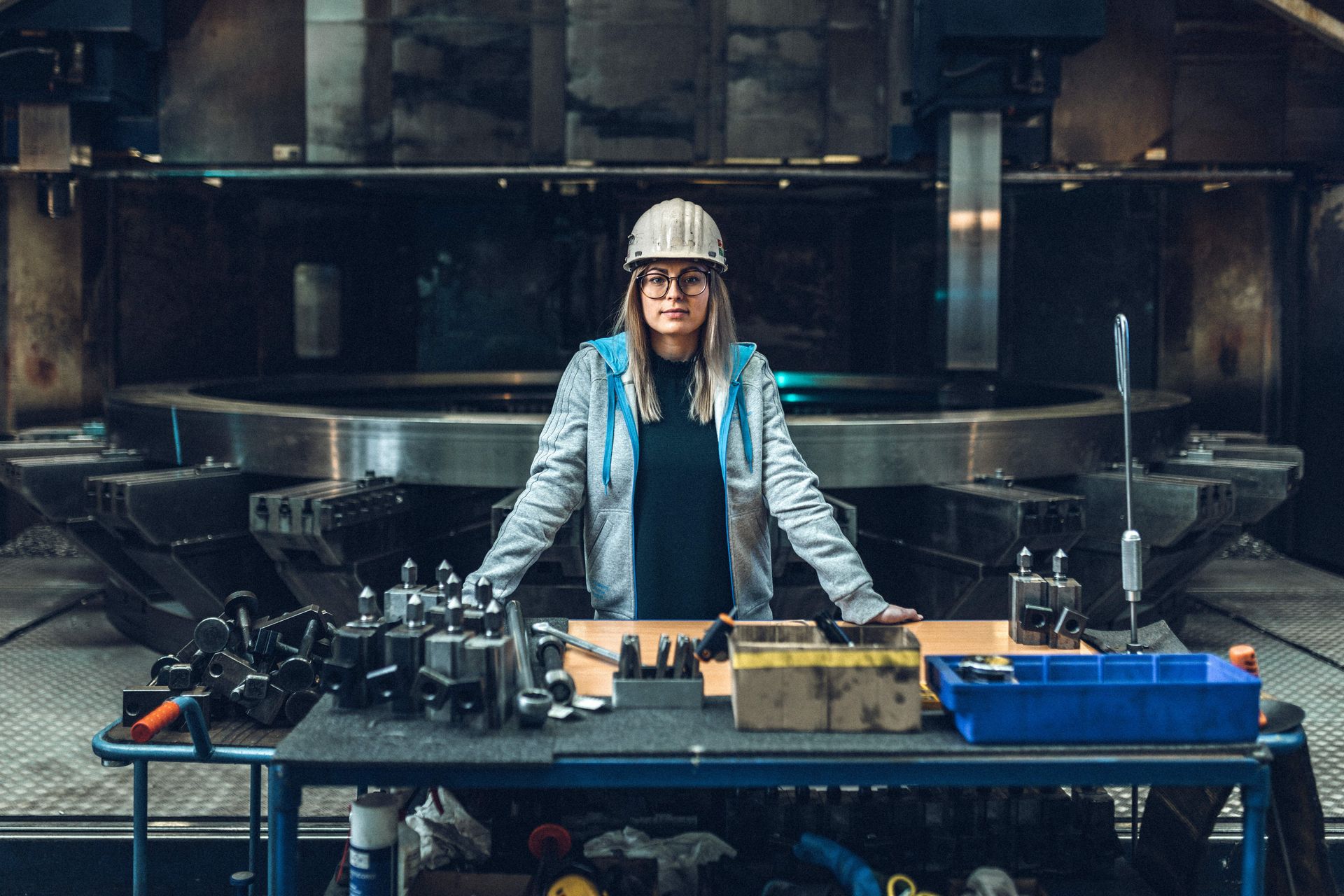 A female employee wearing a hard hat and safety goggles stands at a large workbench in an industrial production hall at thyssenkrupp. Various tools and technical components are laid out on the table. Machines and equipment can be seen in the background. The image conveys a modern, hands-on working environment and highlights thyssenkrupp’s technical expertise.