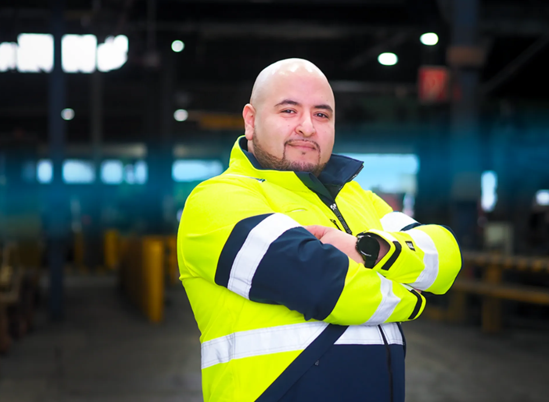 männlich gelesene Person mit verschränkten armen, trägt eine gelbe Schutzjacke, schmale Bartfrisur