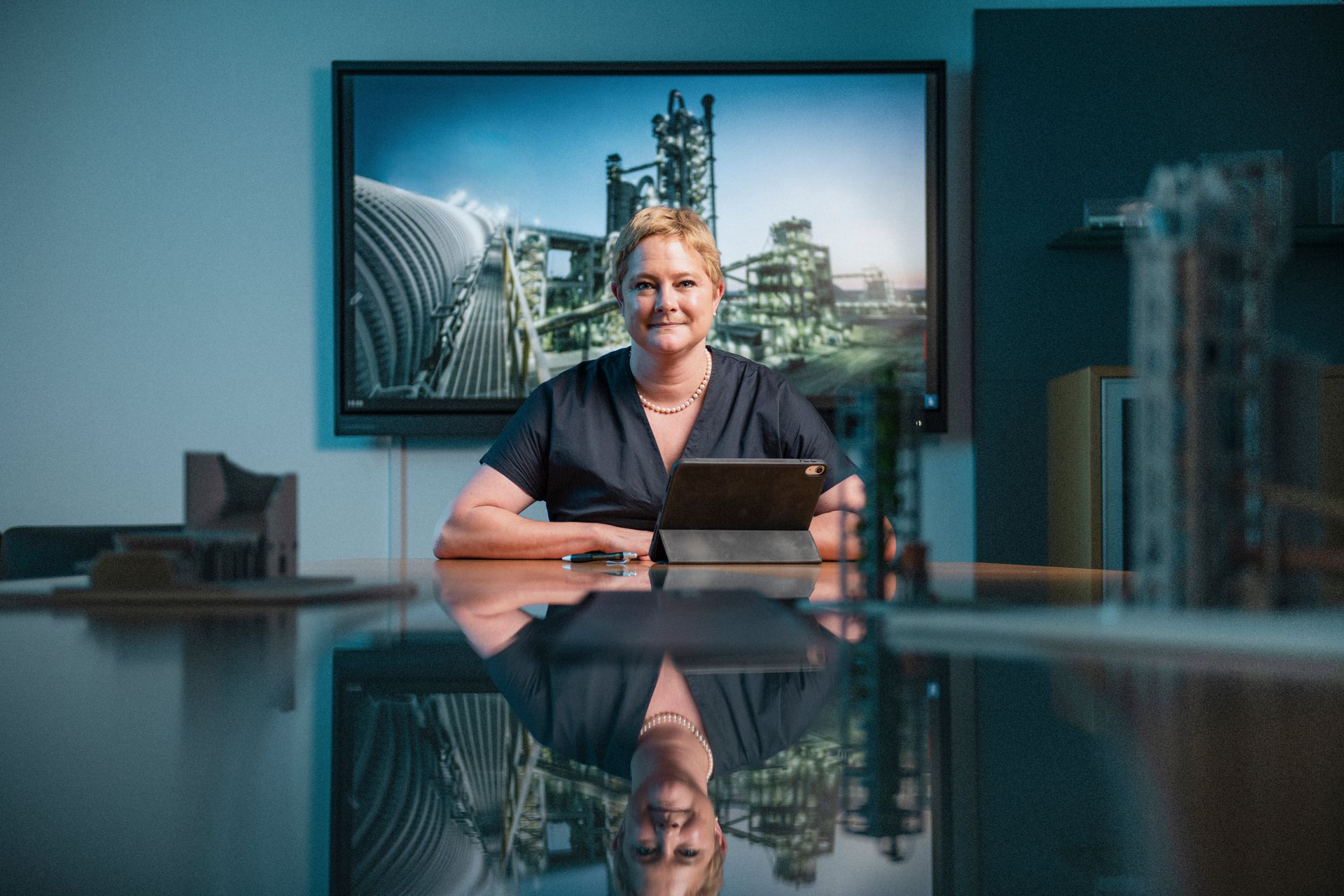 Eine Frau sitzt an einem Konferenztisch und nutzt ein Tablet. Im Hintergrund hängt ein großes Bild einer Industrieanlage. Auf dem Tisch spiegeln sich die Szene und Modellstrukturen.
