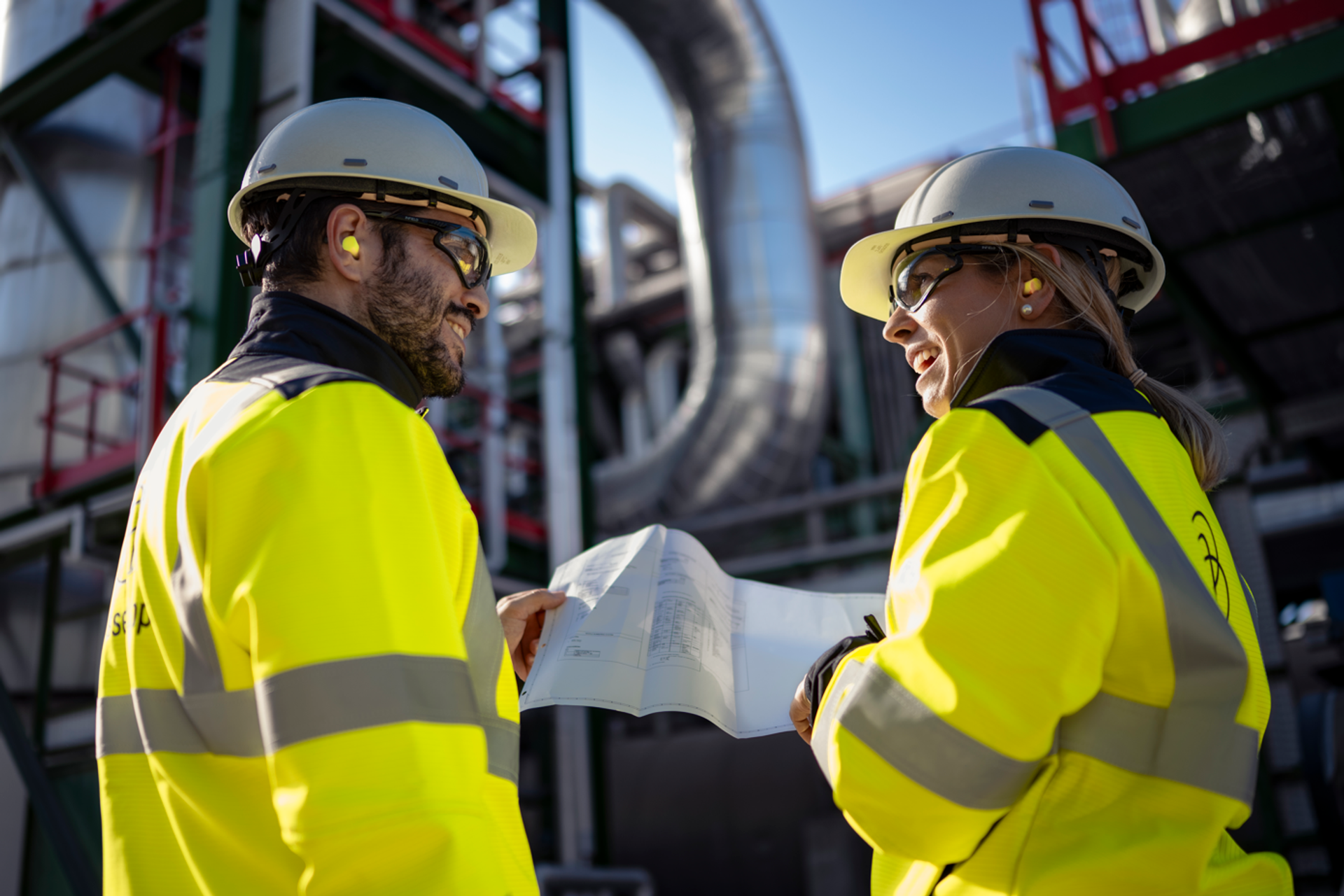 Zwei Mitarbeitende in Sicherheitskleidung und mit Helm besprechen technische Unterlagen auf einem Industriegelände.