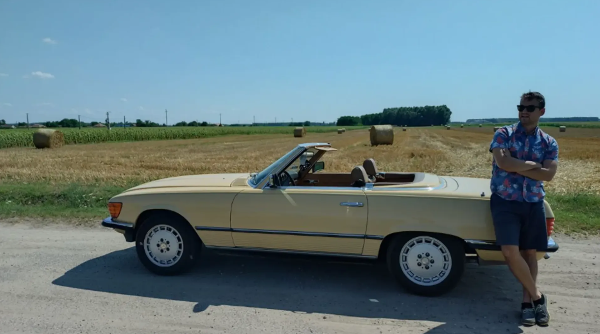 Male-presenting person wearing sunglasses leans with arms crossed against a beige convertible in front of a field with hay bales.
