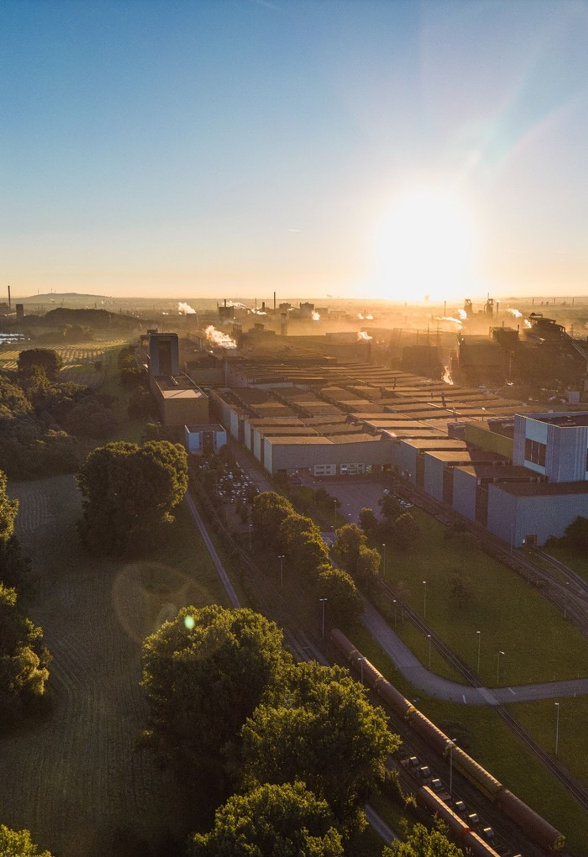 Luftaufnahme des thyssenkrupp-Steel-Werks bei Sonnenuntergang mit Industrieanlagen, Gleisen und Grünflächen.