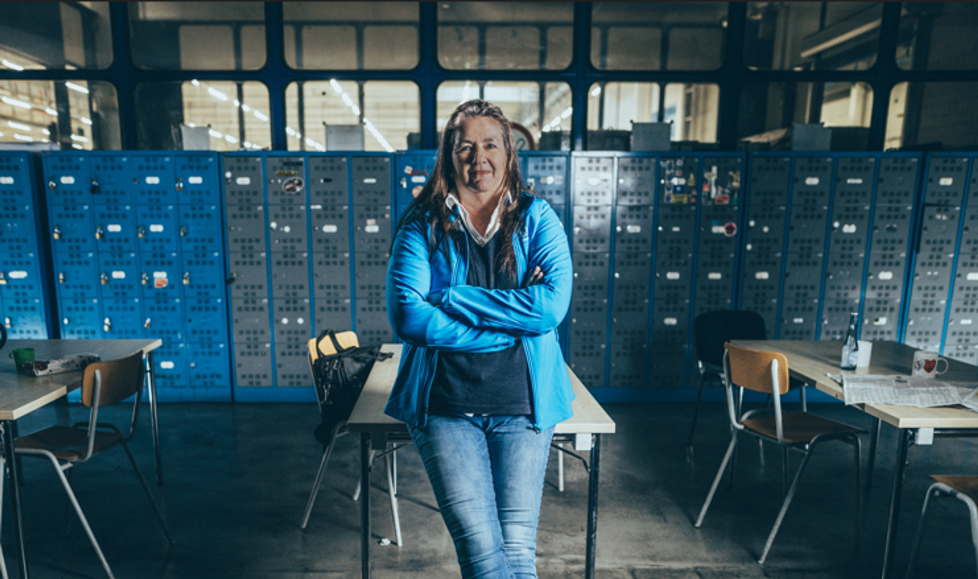 female person wearing a blue jacket, leaning against a desk with arms folded and looking into the camera.