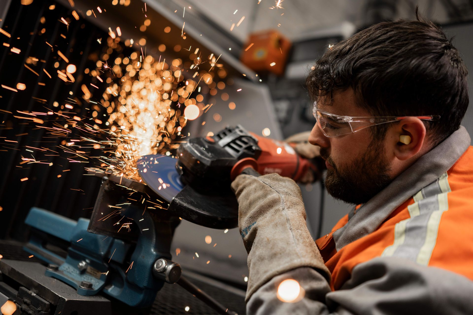 Funken sprühen beim Schleifen eines Metallstücks in einer Werkstatt. Die Person trägt Schutzkleidung und Gehörschutz.