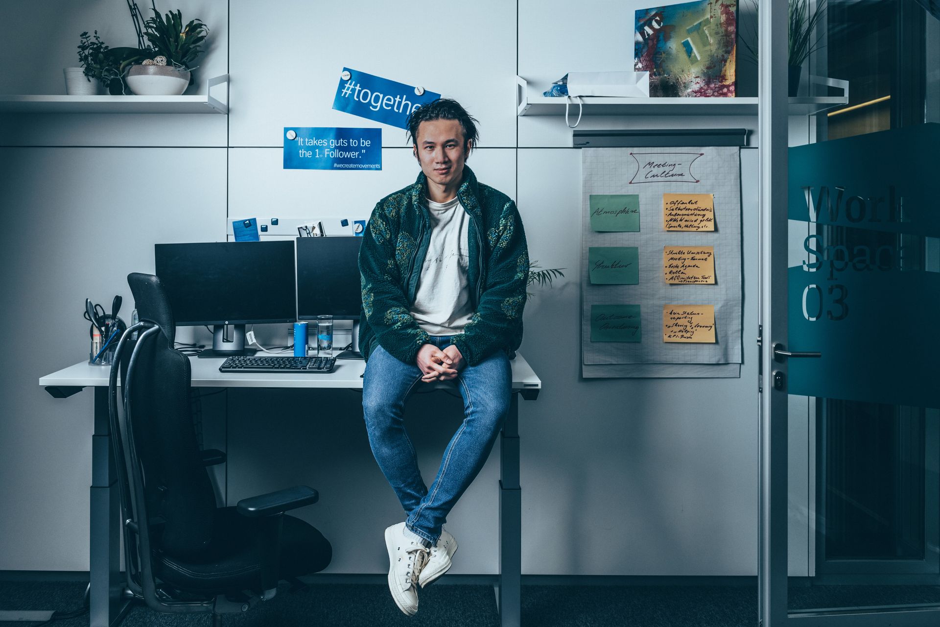 Mann sitzt auf einem Schreibtisch in einem modernen Büro, neben zwei Monitoren und einer Pinnwand mit Notizen.