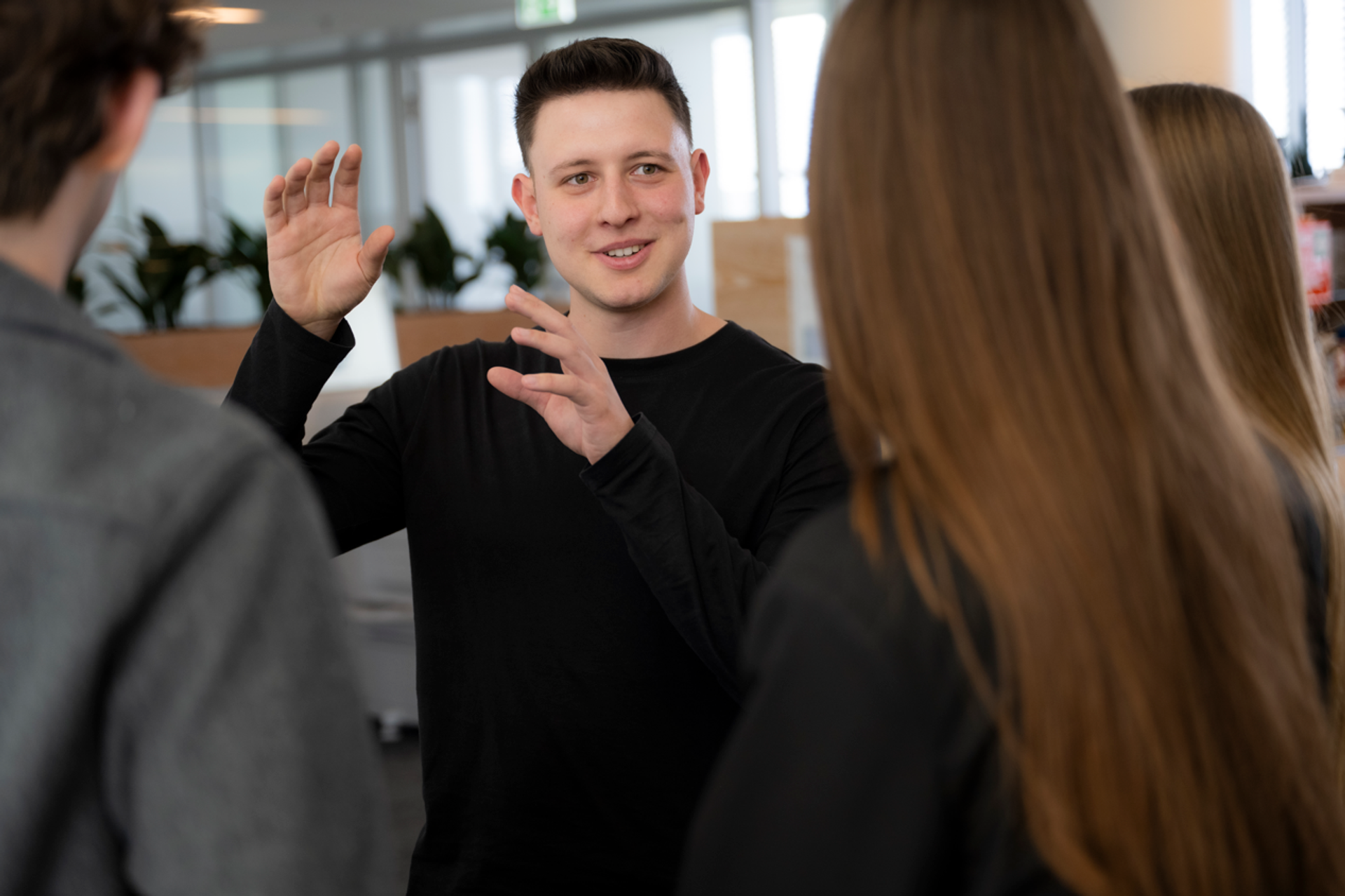 Person erklärt etwas in einer Gesprächsrunde im offenen Bürobereich, umgeben von weiteren Personen.