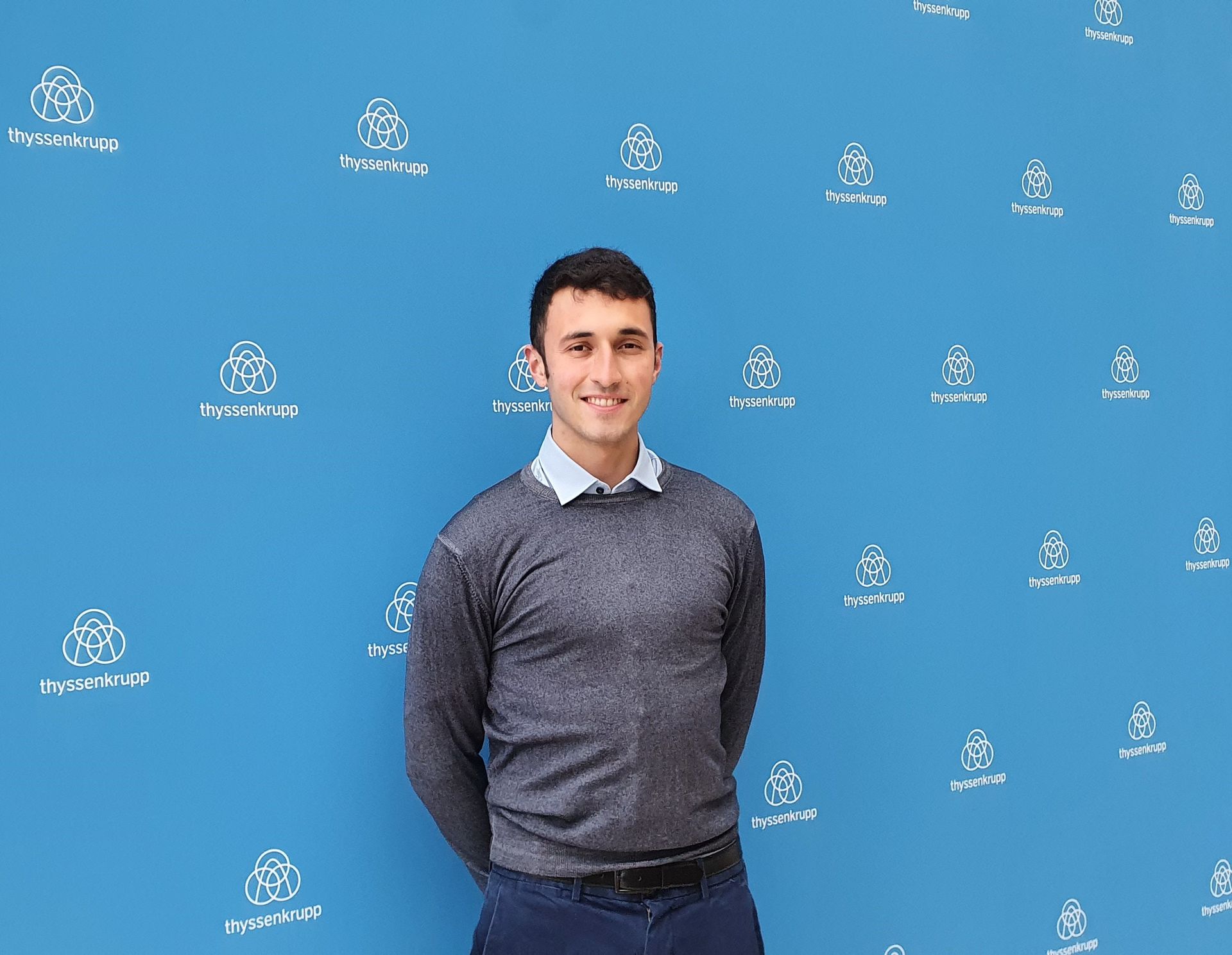 Sebastian Dirk Weidenbach standing in front of a blue corporate background with company logos, wearing business casual clothing.