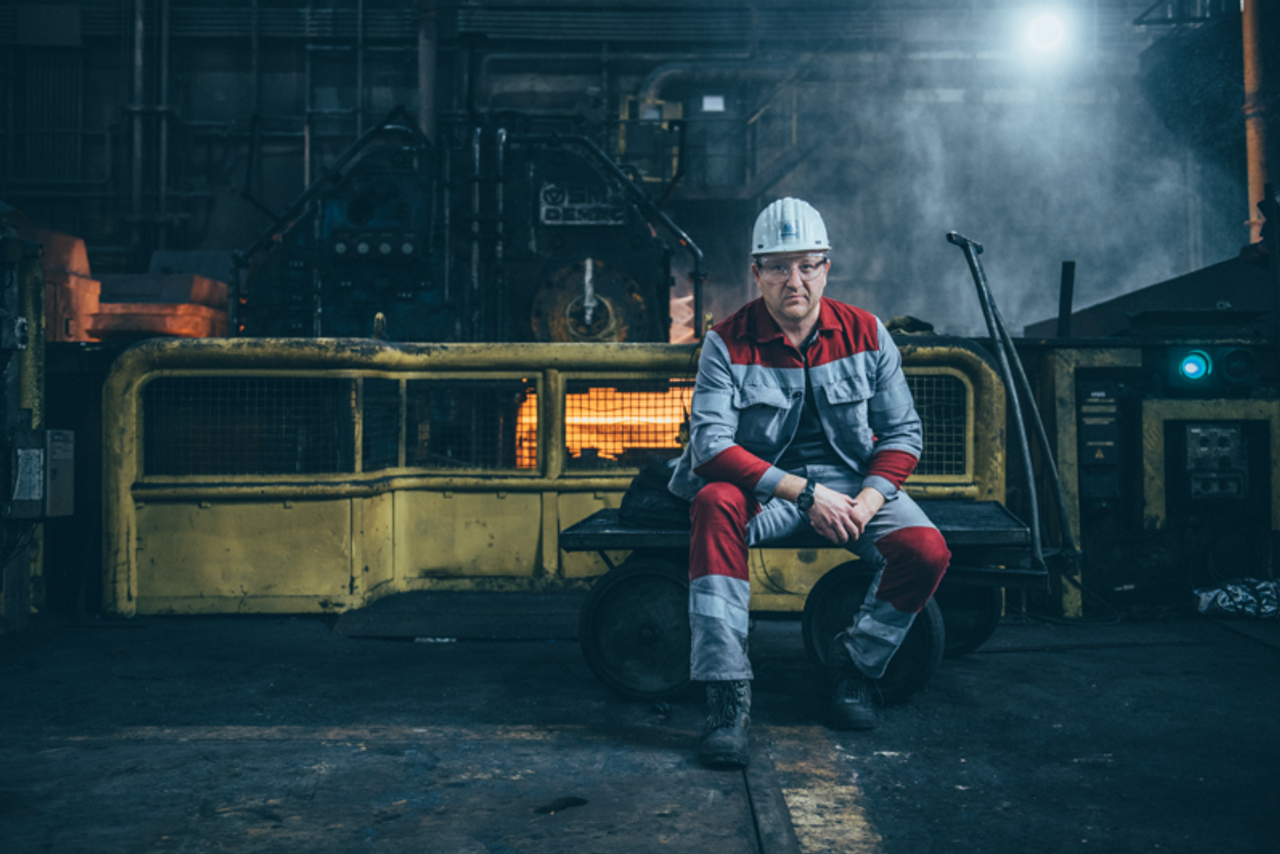 Industriearbeiter in roter und grauer Schutzkleidung sitzt mit Helm in einer Produktionshalle vor glühendem Ofen und schwerem Maschinenbau.
