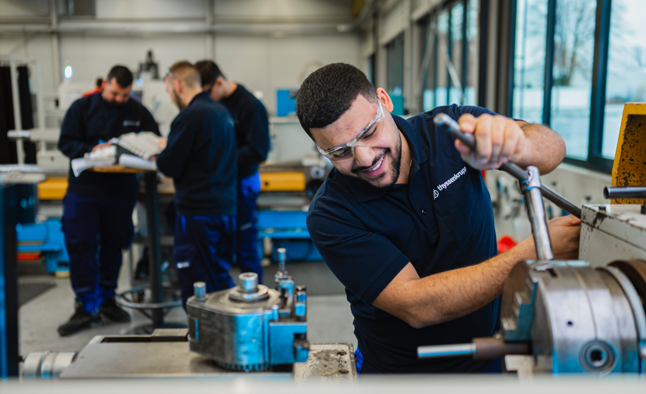 Smiling young man working on a metal machine in a bright, modern workshop. In the background, several colleagues are focused on technical tasks.